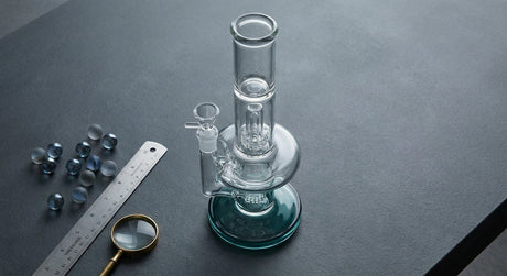 A clear glass bong displayed on a clean surface alongside a magnifying glass and glass marbles, styled in a crisp editorial flat-lay composition with cool blue and charcoal tones.