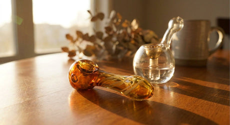 A glass hand pipe and a bubbler resting side by side on a wooden surface in warm natural light, suggesting an everyday carry comparison.
