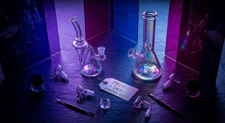 A curated flat-lay of premium glass dab rigs and bongs dramatically lit with purple and blue lighting on a dark slate surface.