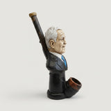A sculptural smoking pipe shaped like a bust portrait of a person in profile, with a long dark stem and a brown bowl, displayed on a white tabletop near a window with an outdoor city view.