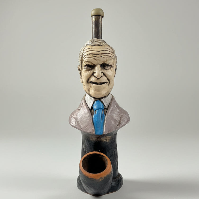A ceramic figurine of a man's bust wearing a suit and blue tie, with a smoking pipe shaped like his head and neck, displayed on a white table in front of a window.