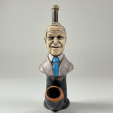 A ceramic figurine of a man's bust wearing a suit and blue tie, with a smoking pipe shaped like his head and neck, displayed on a white table in front of a window.