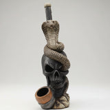 A decorative skull sculpture with a coiled snake draped over it and a small bowl or cup in front, displayed on a white tabletop near a large window with an outdoor city view.