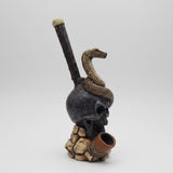 A decorative smoking pipe sculpture displayed on a white table in front of a window. The pipe features a dark skull base with a coiled snake on top, a long dark stem, and a brown bowl resting on a rock-like textured stand.